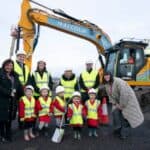 A group of adults and children in safety vests and helmets stand smiling in front of a large yellow excavator at a construction site. The adults wear dark coats; the children wear red outfits and hold small shovels.