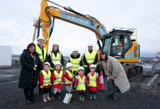 A group of adults and children in safety vests and helmets stand smiling in front of a large yellow excavator at a construction site. The adults wear dark coats; the children wear red outfits and hold small shovels.