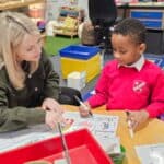 A teacher sits at a round table with a young student in a classroom, helping him with a worksheet. The classroom is colorful and organized, with art supplies and learning materials visible in the background.