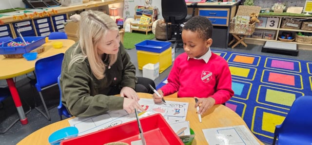 A teacher sits at a round table with a young student in a classroom, helping him with a worksheet. The classroom is colorful and organized, with art supplies and learning materials visible in the background.