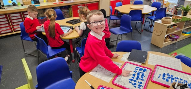 A smiling young boy in glasses, wearing a red sweater, stands at a classroom table with worksheets. Other children in red sweaters work at nearby tables with tablets. The room has blue chairs and colorful decor.