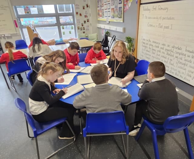 A teacher sits at a round table with six young students, helping them with their writing assignments. Other children work at tables in the classroom, which has large windows and colorful decorations on the walls.