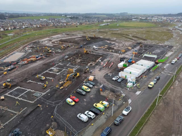 Aerial view of a large construction site with machinery, portable buildings, parked cars, and groundwork in progress, surrounded by roads and fields, with a residential area in the background.