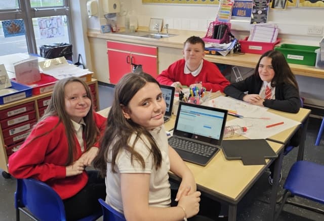 Four students sit around a classroom table covered with papers, pens, and a laptop. Three wear red uniforms, one wears a white shirt. They are smiling and working together on a project. Classroom supplies and posters are in the background.