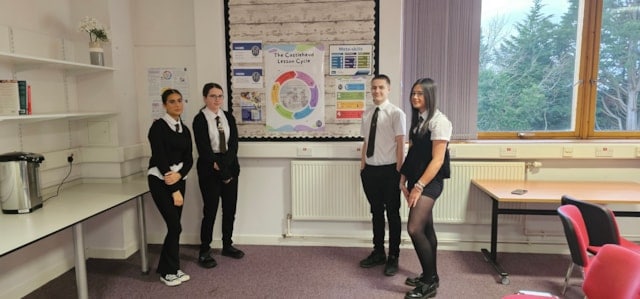 Four students in school uniforms stand in a classroom near a bulletin board displaying a colorful chart. There are shelves, a window with blinds, tables, chairs, and trees visible outside.