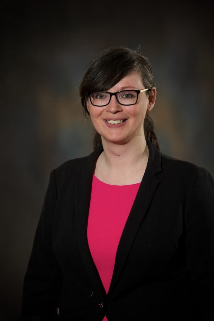A woman with straight dark hair, glasses, and a black blazer over a bright pink top smiles at the camera against a blurred dark background.