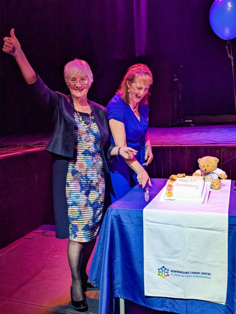Two women stand by a table with a decorated cake and a teddy bear. One woman gives a thumbs up while the other cuts the cake. Both are smiling. The setting appears festive with a purple-lit stage and blue balloons.