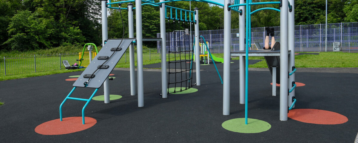 Modern playground structure with climbing bars, rope net, and slide on a rubber surface. A child sits at the top with legs visible, surrounded by green grass and trees in the background.