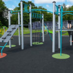 Modern playground structure with climbing bars, rope net, and slide on a rubber surface. A child sits at the top with legs visible, surrounded by green grass and trees in the background.