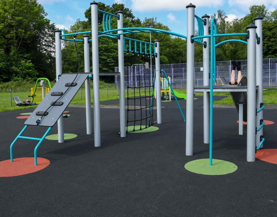 Modern playground structure with climbing bars, rope net, and slide on a rubber surface. A child sits at the top with legs visible, surrounded by green grass and trees in the background.