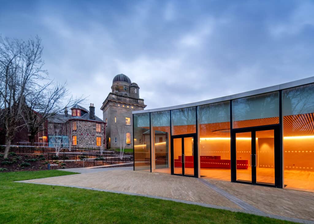 Modern glass building with warm interior lighting stands next to an older stone building with a domed observatory tower, under a cloudy evening sky with green grass in the foreground.