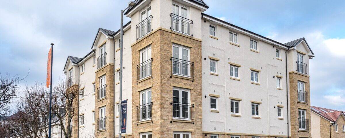 A modern four-story apartment building with light stone and white exterior walls, small balconies, and a dark gray tiled roof, situated on a corner lot with a sidewalk, trees, and a cloudy sky.