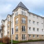 A modern four-story apartment building with light stone and white exterior walls, small balconies, and a dark gray tiled roof, situated on a corner lot with a sidewalk, trees, and a cloudy sky.