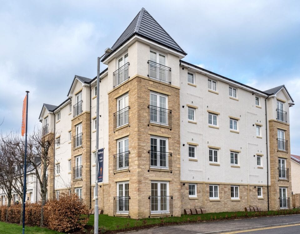 A modern four-story apartment building with light stone and white exterior walls, small balconies, and a dark gray tiled roof, situated on a corner lot with a sidewalk, trees, and a cloudy sky.