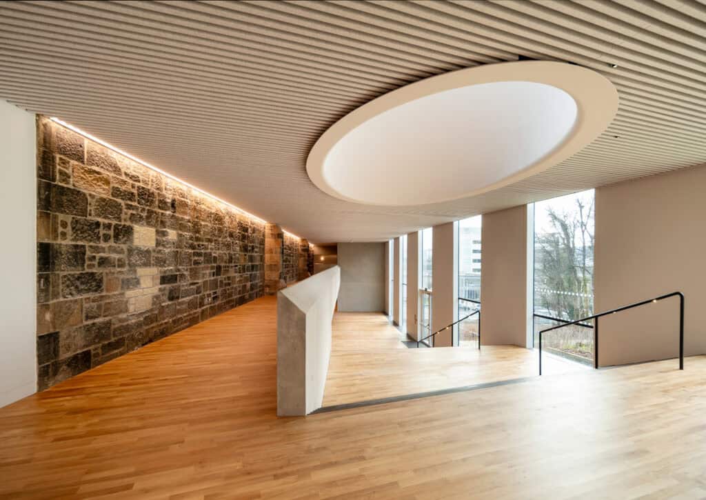 Modern interior with wooden flooring, a stone accent wall, large windows, and a ceiling with a large oval recess. Black handrails line a few steps leading down. Natural light fills the space.
