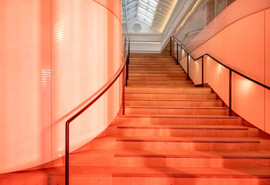 A modern indoor staircase with orange-tinted lighting, sleek black handrails, and a curved wall on the left, leading up to a glass ceiling that lets in natural light.