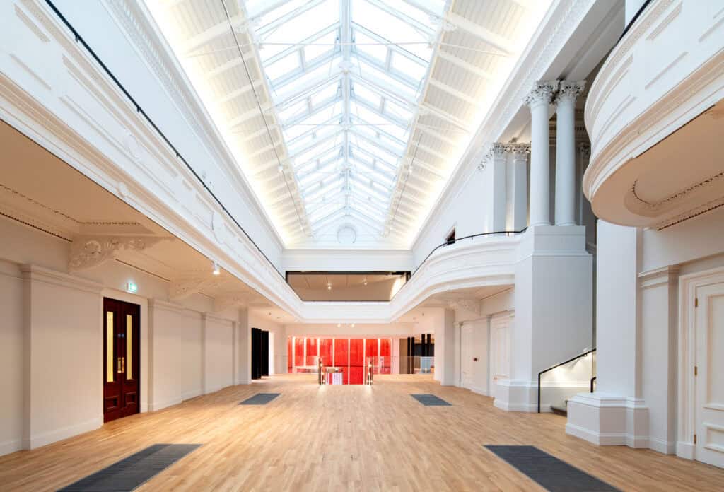 Spacious, bright hall with high glass ceiling, white walls, ornate columns, wooden floors, and a red accent wall at the far end. Modern and classic architectural features blend seamlessly.