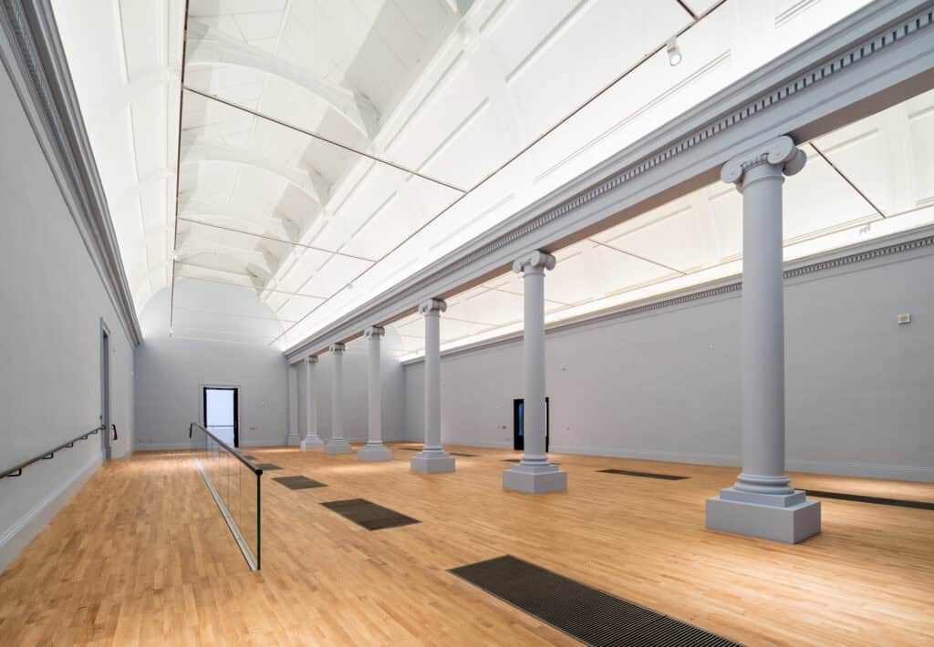 A spacious, empty gallery with high vaulted ceilings, white walls, and tall gray columns. The floor is light wood, and natural light floods the room from skylights above.