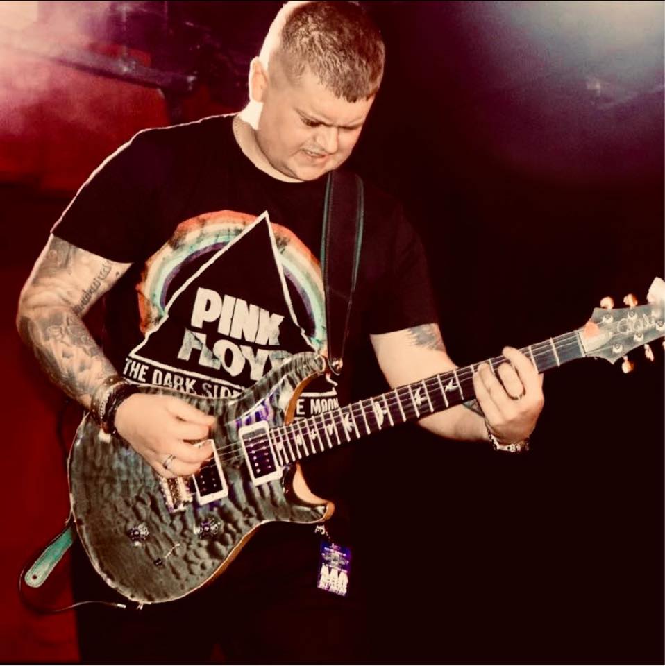 A person with short hair and tattoos plays an electric guitar on stage, wearing a black Pink Floyd t-shirt. The background is dark with red and purple lighting.