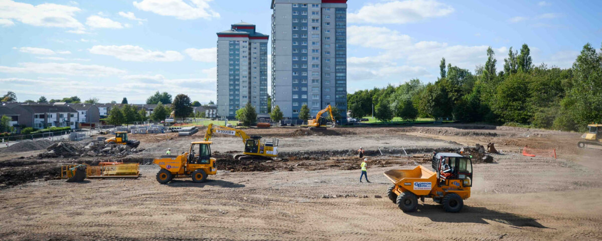 A construction site with several yellow vehicles and workers on dirt ground, with two tall apartment buildings and trees in the background under a partly cloudy sky.