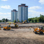 A construction site with several yellow vehicles and workers on dirt ground, with two tall apartment buildings and trees in the background under a partly cloudy sky.