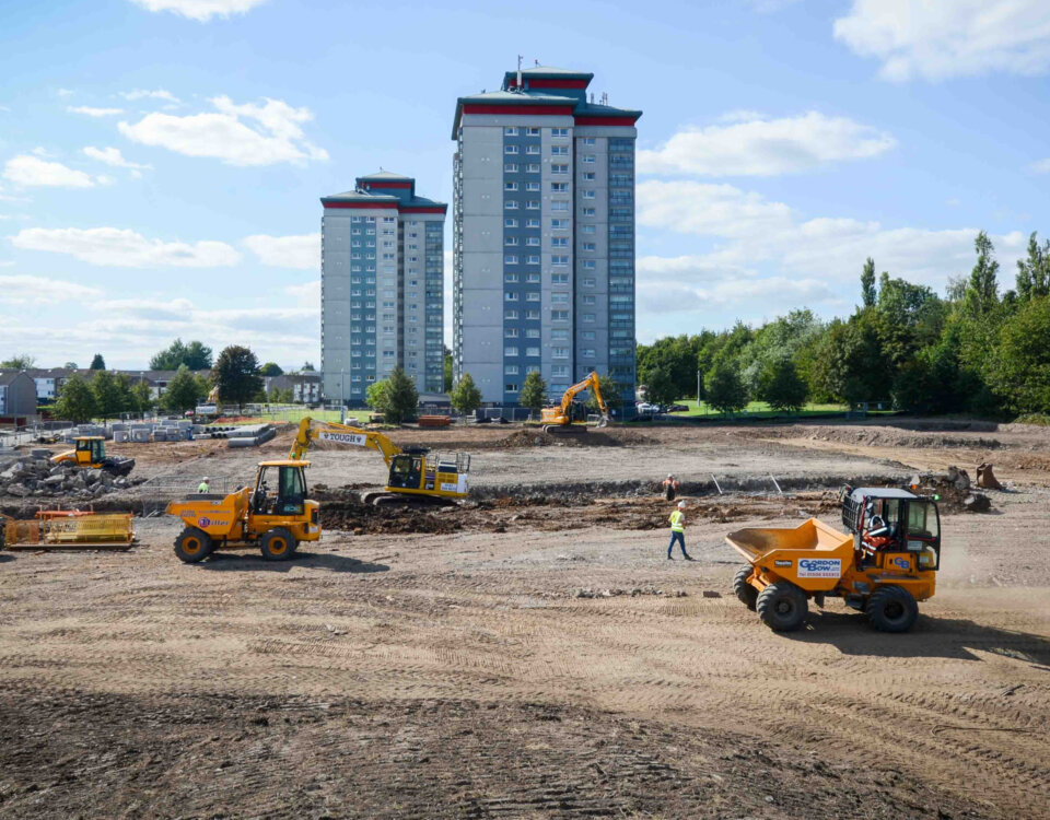 A construction site with several yellow vehicles and workers on dirt ground, with two tall apartment buildings and trees in the background under a partly cloudy sky.
