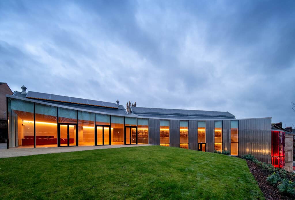 A modern building with large glass windows glows with warm interior lights at dusk. The structure features angular lines and metal cladding, set against a cloudy sky and surrounded by green grass.