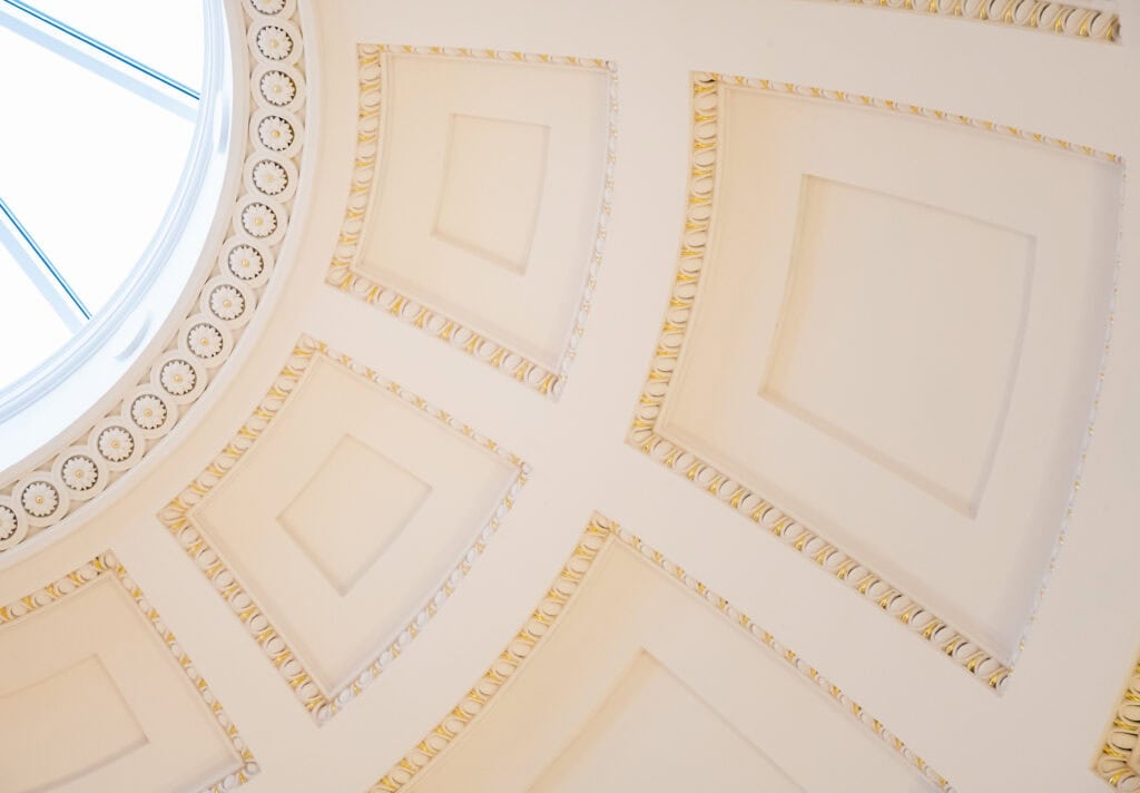 Ornate cream-colored ceiling with decorative square patterns and gold detailing, featuring a circular skylight with sections of glass letting in natural light.