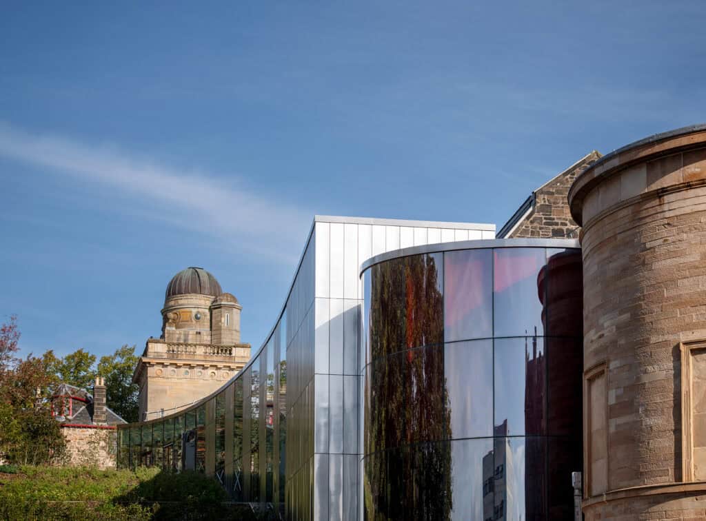 A modern glass building with curved walls stands between two older stone buildings. An observatory dome is visible in the background under a blue sky. Trees are reflected in the glass facade.