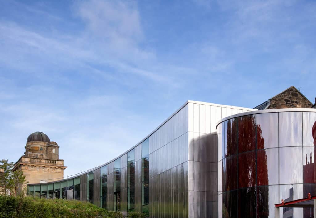 A modern glass and metal building with curved architecture stands beside an older stone building with a domed observatory under a bright blue sky.