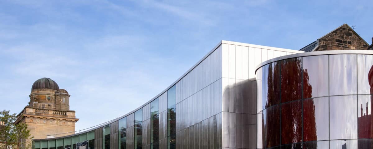 A modern glass and metal building with curved walls stands beside an older stone structure with a dome, against a blue sky.