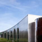 A modern glass and metal building with curved walls stands beside an older stone structure with a dome, against a blue sky.