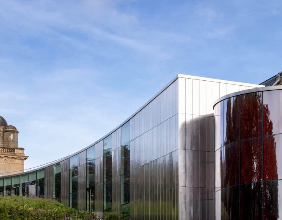 A modern glass and metal building with curved walls stands beside an older stone structure with a dome, against a blue sky.