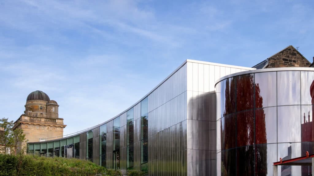 A modern glass building with curved architecture stands beside an older stone structure with a domed observatory under a bright blue sky.
