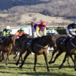 Five jockeys on horses race across a grassy track, each wearing colorful racing silks and helmets, with countryside hills and trees visible in the background.