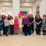A group of 18 adults stand indoors, smiling for a group photo. They are dressed in a variety of formal and semi-formal outfits, with pink balloons in the background and a white column centered behind them.