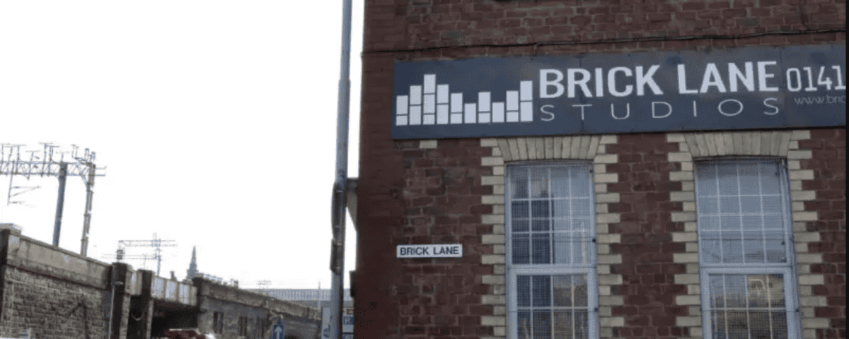 A brick building with a "BRICK LANE STUDIOS" sign faces a busy street with cars, a fire truck, and a bus driving past. A small "Brick Lane" street sign is mounted on the corner of the building.