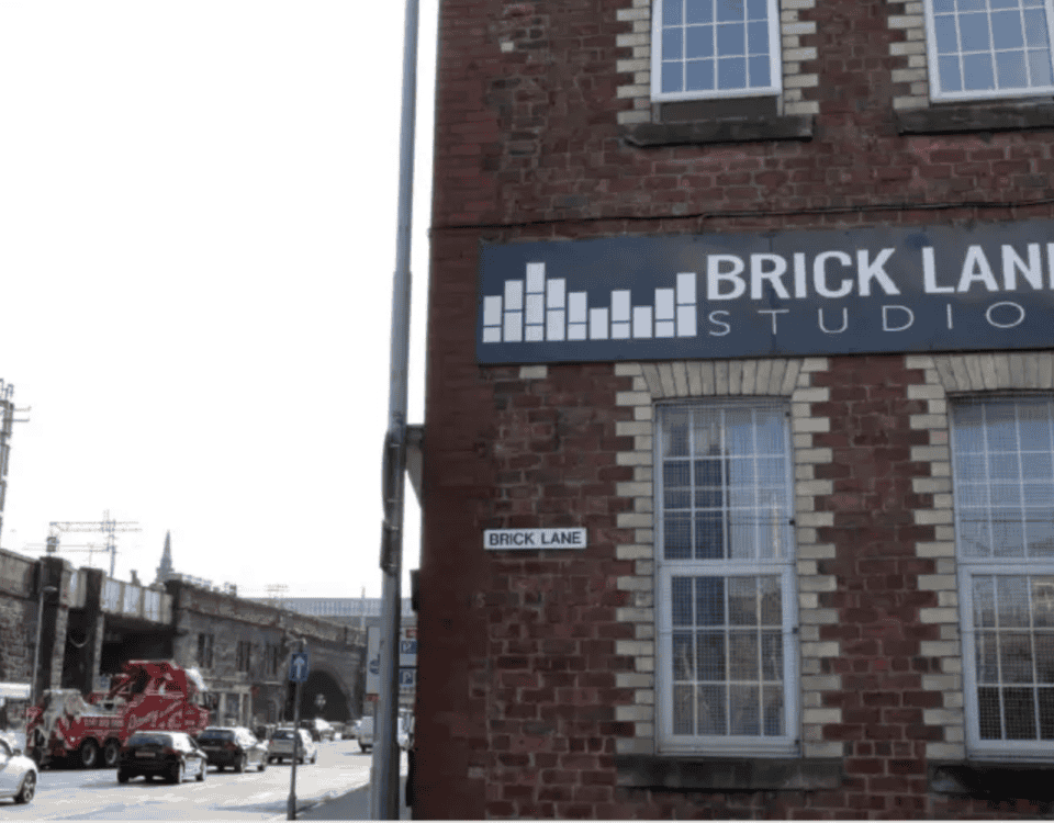 A brick building with a "BRICK LANE STUDIOS" sign faces a busy street with cars, a fire truck, and a bus driving past. A small "Brick Lane" street sign is mounted on the corner of the building.