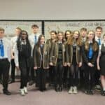 A group of students in uniforms stand indoors with a teacher. Most wear blazers and ties, and they are smiling at the camera. They stand in front of a bulletin board that reads "VISIBLE LEARNING.