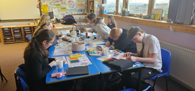 A group of students sit around a table in a classroom, focused on drawing and painting with art supplies. Papers, paint sets, and brushes are scattered across the table. Colorful posters and large windows line the walls.