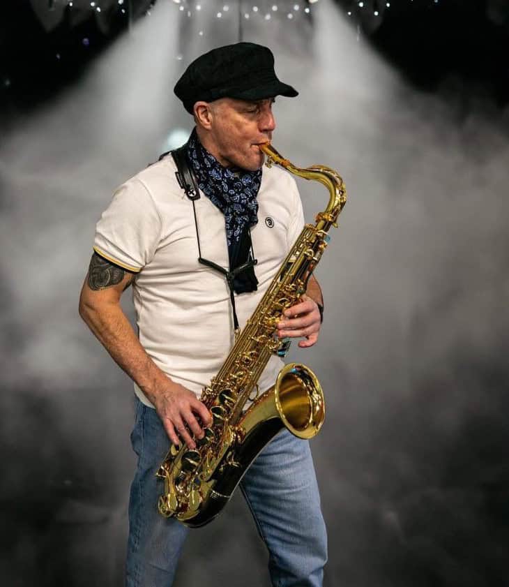A man wearing a black cap, patterned scarf, white shirt, and jeans plays a gold saxophone onstage, surrounded by smoke and dramatic lighting.