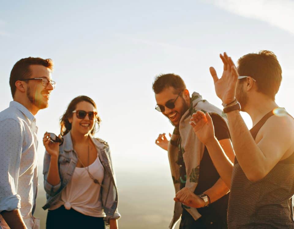 Four people stand outdoors, smiling and laughing together in warm sunlight. Two men wear sunglasses, and a woman holds sunglasses in her hand. The group appears to be enjoying a fun, casual moment.