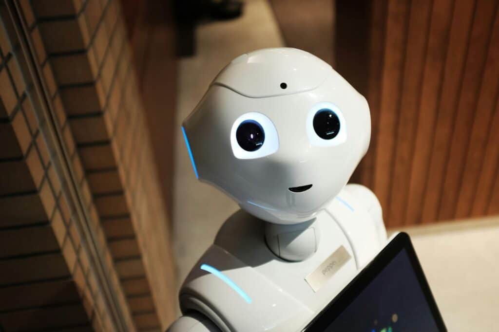 A white humanoid robot with large, expressive eyes and a digital tablet on its chest stands indoors, looking up with a friendly expression. The setting includes brick and wood-paneled walls.