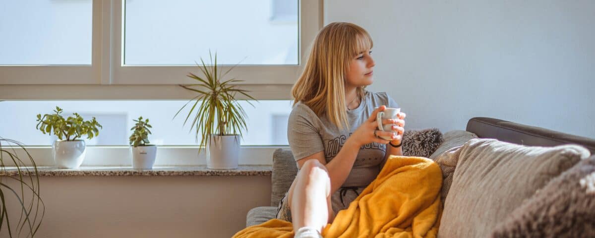 A woman with blonde hair sits on a couch covered with a yellow blanket, holding a mug and looking out a window. There are potted plants on the windowsill behind her, and sunlight fills the cozy room.