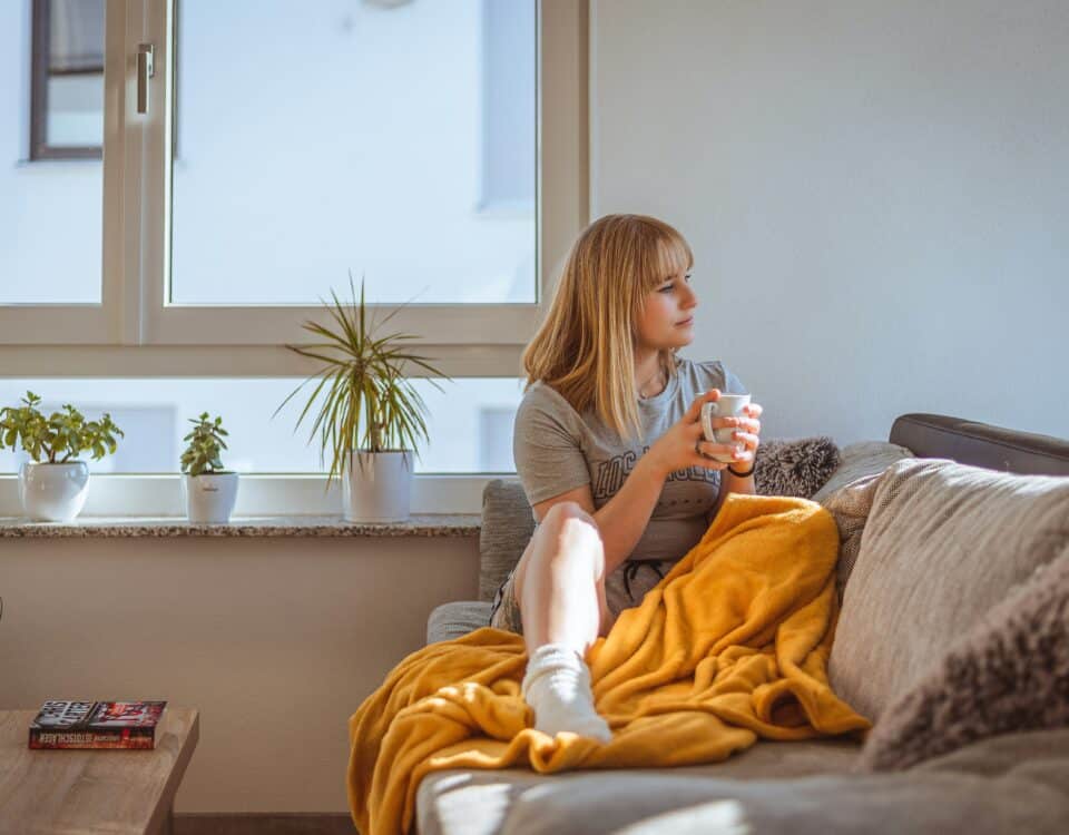 A woman with blonde hair sits on a couch covered with a yellow blanket, holding a mug and looking out a window. There are potted plants on the windowsill behind her, and sunlight fills the cozy room.