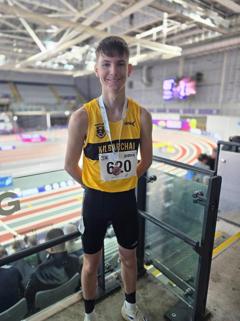 A young male athlete in a yellow "Kilbarchan" running vest and black shorts stands indoors, smiling with a medal around his neck and a race number (620) pinned to his shirt. Indoor athletics track visible in the background.