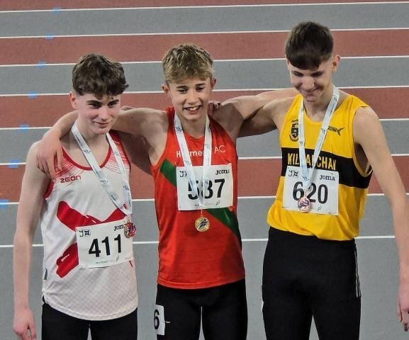 Three young male athletes stand on a podium, smiling with arms around each other. They wear medals and race bibs numbered 411, 387, and 620. A photographer kneels in front to take their photo on an indoor track.