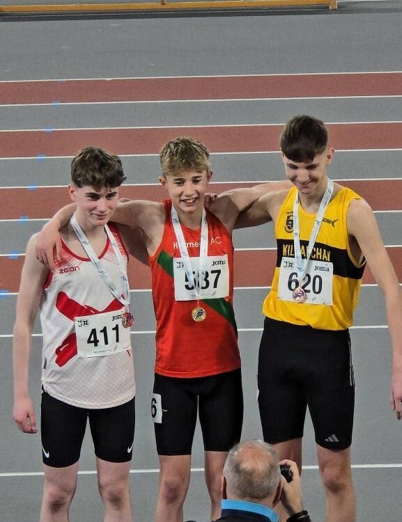 Three young male athletes stand on a podium, smiling with arms around each other. They wear medals and race bibs numbered 411, 387, and 620. A photographer kneels in front to take their photo on an indoor track.