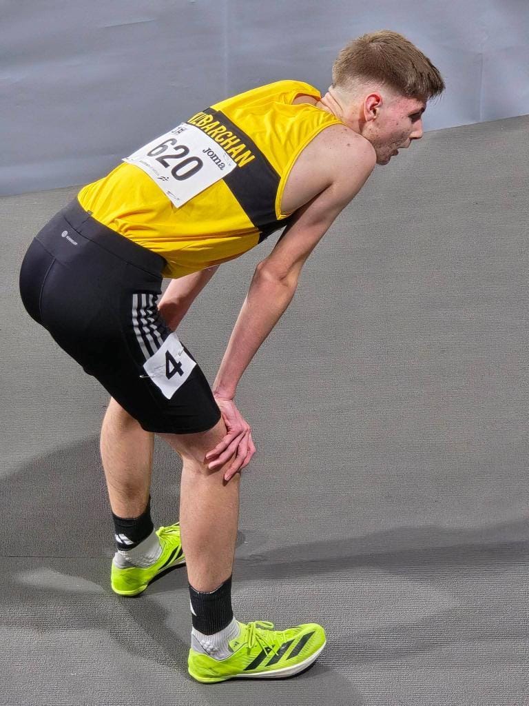 A male athlete in a yellow tank top and black shorts stands bent over with hands on knees, appearing fatigued after a race. He wears bright green running shoes and a race bib numbered 620.