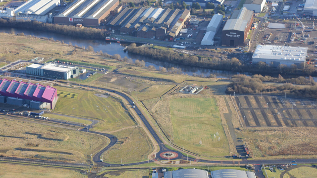 Aerial view of an industrial area with warehouses, a river, fields, roads, a roundabout, and scattered buildings; some grassy and open spaces are visible alongside the developed land.